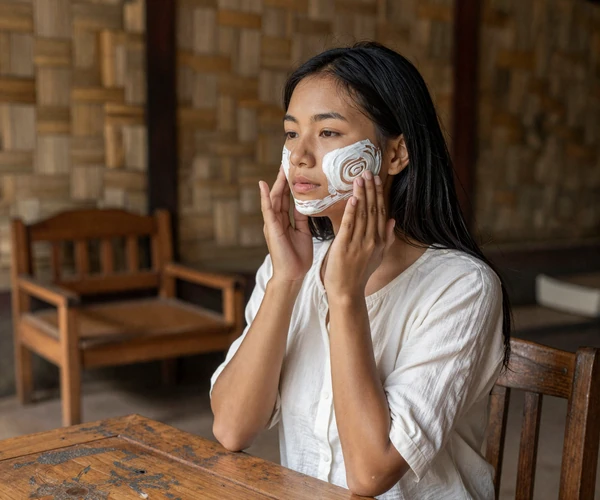 Young Burmese woman applying traditional thanaka paste on her cheek in a traditional Myanmar home